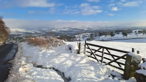 BBC Weather Watchers/Ganty A view of snow-covered fields stretches to the horizon. A field gate is on the right with a road on the left.