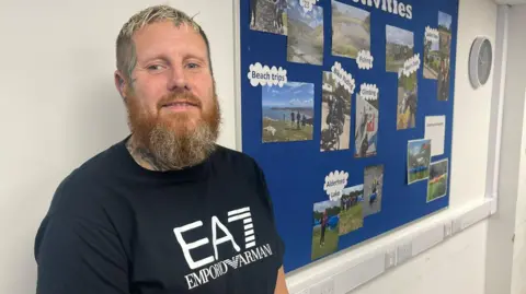 A man with short bleached hair and a black t-shirt with large centre logo stands in front of a pinboard on a white wall. The blue pinboard has photographs of people taking part in outdoor activities. The man is smiling through his bushy ginger beard. He has tattoos on his neck and the side of his face.