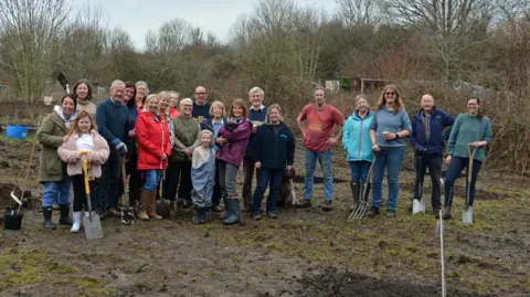 Peter Thompson Group of people smiling widely together on a muddy surface outdoors. Some hold spades and trowels.