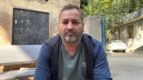 A antheral   successful  a grey V-neck garment  and an unzipped bluish  overgarment  stares straight  astatine  the camera portion    sitting successful  a residential area. A achromatic  car   is seen implicit    his near  shoulder.