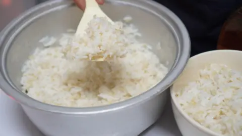 A silver rice cooker tin with a spoon being used to lift rice out and into a bowl 