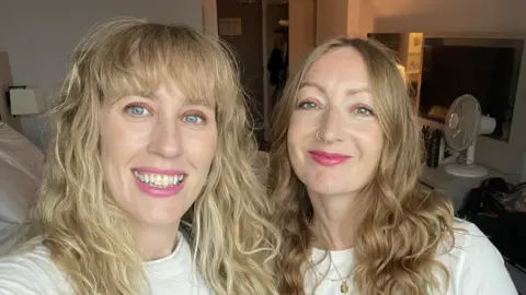 Nicola Brown Nicola and Lowri. Both have long wavy hair and are wearing pink lipstick and white t-shirts. 