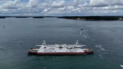 Image shows an aerial shot of the Sandbanks chain ferry. The ferry has a red base with the rest of the sides white. Behind the ferry, you can see land. 
