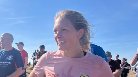 Connor Bennett/BBC A young woman with a pink running top on smiling while looking away from the camera. She is at the start line of a park run on a beach and there are other runners behind her.