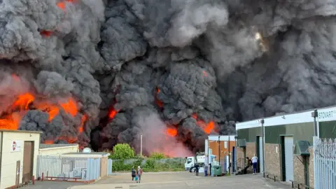 Huge cloud of dark grey smoke billowing from the ground, with flame visible in the lower half. There are bushes in front of the smoke. There is a single-storey building to the right and another to the left. There are people standing on the yard in between.
