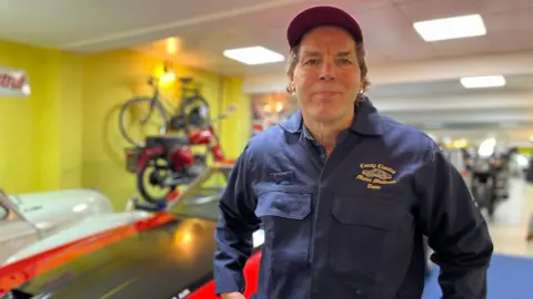 Pat wearing a red trucker cap and dark blue overalls standing in his museum. A car and a motorcycle can be seen behind him but out of focus.