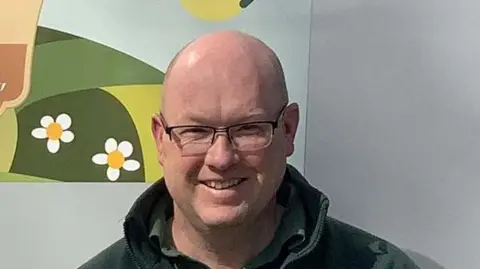 The Rural Coffee Caravan Garry Simmonds is smiling at the camera. He is balding and is wearing spectacles and a green top.