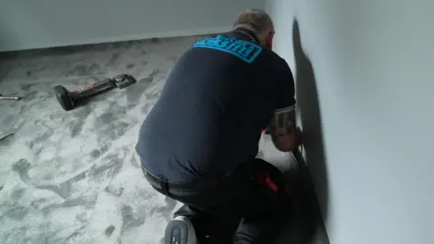 Un hombre coloca una alfombra gris en una habitación vacía, con herramientas a su lado.