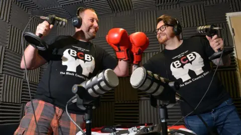 Kevin Dillon (left) and Lee Cadman (right) in the Radio WM studio talking about mental health on Ed James's Mid Morning programme. The pair have microphones, headphones and other radio equipment around them and they are both wearing a boxing glove and smiling at each other.