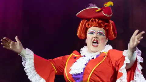 A pantomime dame on stage, with their arms outstretched and their mouth open. The dame has bright-red lipstick on and is wearing a pirate's hat with a parrot on it, a bright red wig, and a red dress with white frills. 