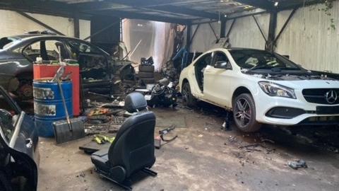 A chop shop, with two stripped down cars, including a white Mercedes