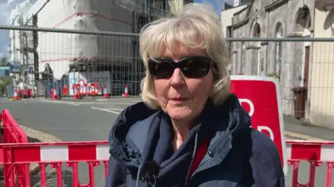 The image shows a person standing in the foreground behind red and white temporary road barriers. The person is wearing a dark padded gilet over a long‑sleeved top and has a red lanyard around their neck. A small microphone is clipped to their clothing, suggesting they are being interviewed on camera.