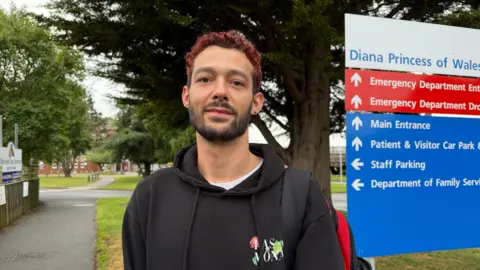 Christian Wiltshire has red hair and is wearing a black hoodie. He is stood close to a sign for Diana, Princess of Wales Hospital.