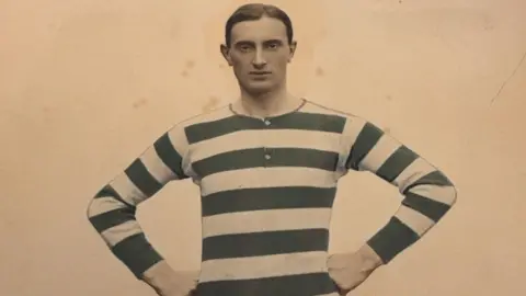 A footballer pictured in the early 20th century. His football shirt is striped. He has short, smart hair and a neutral expression. 