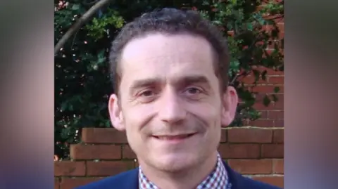 Ian Fisher smiles at the camera. He has short curled dark hair. He wears a blue suit with a blue, red and white chequered shirt underneath. A red brick wall as well as a holly bush can be seen behind him.