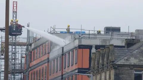 Joshua Brandwood Firefighters on an aerial platform use water pumps to put out a blaze at the top of a block of flats under construction in Lancaster