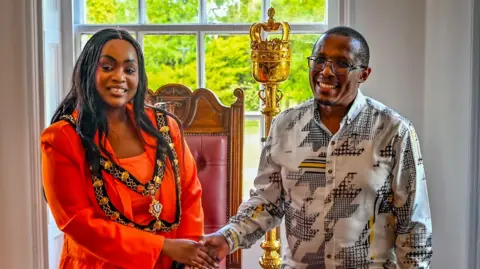 John Chatterley A black woman dressed in red, with chains around her shoulders, shaking hands with a black man with glasses. There is a mace in the background and a window looking out on to some trees