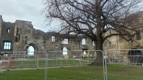 Interior of castle showing grassy area criss-crossed with safety fencing and surrounded by a ruined castle wall with sky visible through windows