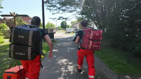 A white woman and white man, walking up a path with grass and trees either side, both wearing black t-shirts and orange trousers, wearing backpacks and carrying bags in their hands, approaching a street with several houses