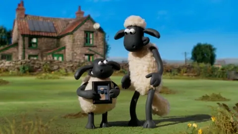Two black and white, clay animation sheep are standing on grass in front of a farmhouse-style cottage.