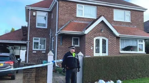 A police officer stands outside a house with police tape across the front of it