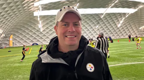 Rooney is wearing a white gap and black jacket as he smiles at the camera in front of kids playing sports.