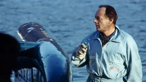 Getty Images Donald Campbell smokes a cigar as he stands next to Bluebird in January 1967. He has dark hair brushed back from his face and is wearing blue overalls.