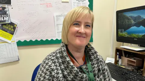 Ollie Conopo/BBC A woman in a black and white jumper sits at her desk looking at the camera.