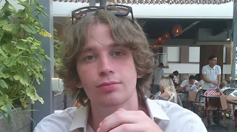 Linus Rowland-Bell A young man with brown, centre-parted hair sits at a table in an outdoor dining area, sunglasses resting on top of his head. Behind him other guests can be seen as waiters take orders and clear tables.