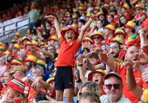 Getty Images A boy stands over his seat at the stadium, cheering for Wales with his arms in the air. He is wearing a bucket hat and the Welsh jersey