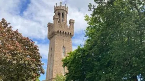 Guernsey's Victoria Tower is seen through the trees while the sky in the background is blue with a lot of cloud coverage