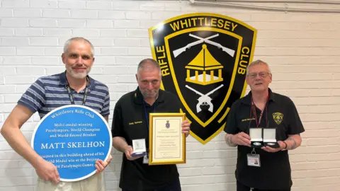 Tom Jackson/BBC The three men are standing against a white bricked wall with the Whittlesey Rifle Club logo on it. They are holding medals and certificates. One is a large blue plaque noting that the paralympian Matt Skelhon trained at the club.