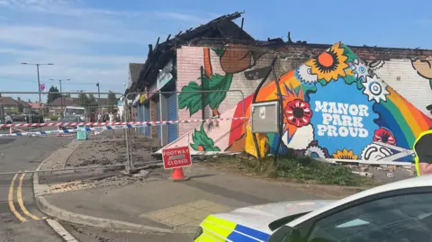 Simon Thake/BBC Tiles and bricks from the affected buildings litter the pavement at Manor Park Centre, off Harborough Road. Part of a mural is visible on the left hand side. The shops all have blue shutters down. A fence is up with police tape across it.