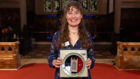 Andy Sillett Libby Watts, who has long brown hair and is wearing a blue dress, stands up while holding her framed architecture prize, inside a church. An alter and stain glass windows can be seen in the background.