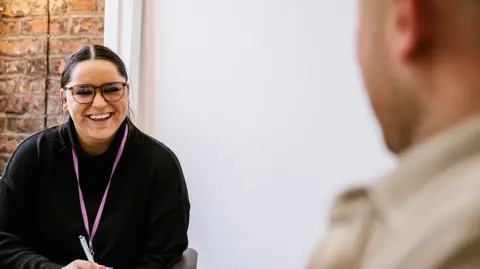 A staff member at the rehabilitation site with brown hair and brown glasses wearing a black top sits in front of a desk during a session with man who has his back to the camera and is blurred out.