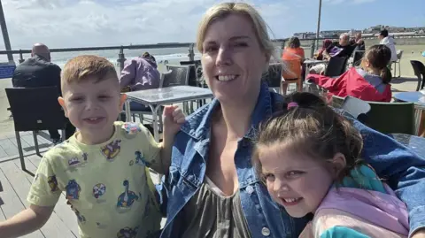 Stacey Angel Photograph of Stacey Angel (Centre) and her son, Brayen, (left) and her granddaughter. Stacey has blonde hair and smiles at the camera. They all sit on a patio looking out over the seaside. 