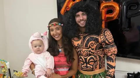 Morgan Deakin A family of three in fancy dress standing in front of a television with large orange balloons behind them
