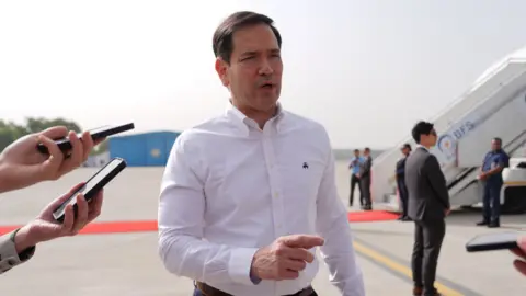 Marco Rubio in a white shirt, pointing outside a plane on a runway in New Delhi