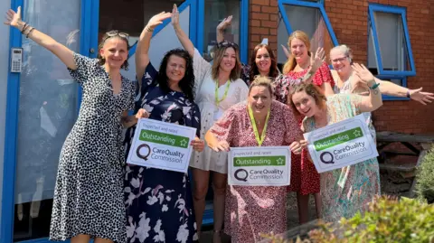 Home Instead A large group of women celebrating, with their arms out and holding signs. They are all in front of a building. 