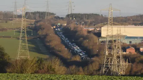 RSM Photography A aerial view of the A1 close to the scene of a crash which killed a baby boy. A long queue of traffic is seen backing up into the distance. The carriageway is surrounded by large trees and bushes. Green fields and electrical pylons are also visible.