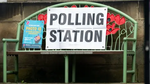 EPA A poster reminds voters to bring their photo ID. It is tied to a green metal bench next to a polling station sign. 