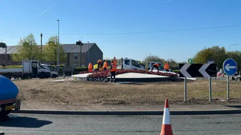 Workmen in orange hi-vis vests are pouring white gravel and planting red flowers into a raised bed in the middle of the roundabout. It is circular and taking the pattern of the St George's cross. Traffic cones have been placed around the inner lane of the roundabout.