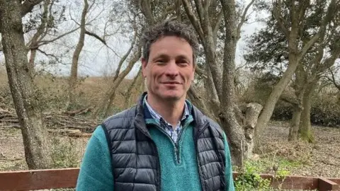 Greg Morel wearing a teal pullover and a dark padded vest is standing outdoors in front of a wooden fence. Behind them is a natural landscape with leafless trees, scattered branches, and open ground, suggesting a woodland or rural area in late autumn or winter.