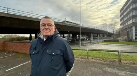 Martin Gannon standing in a car park near the Gateshead flyover. He has short hair and is wearing glasses and a navy coat. The concrete bridge is behind him. Temporary fencing has been put up across the road running under the bridge.
