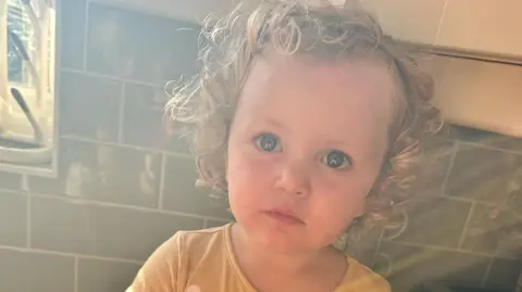 Family handout Amber looks at the camera with wide eyes and a serious expression. She has very curly blonde hair and wears a bright yellow top. She appears to be sitting on a kitchen counter, with green tiles behind her.