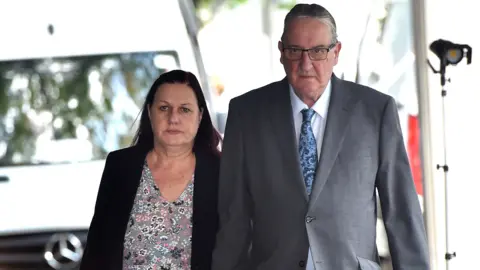 PA Media Susan Letby, who has dyed red hair and a wears a dark blazer over a flowery blouse, and John Letby, who has short grey hair and wears a light grey suit, walk towards Manchester Crown Court hand in hand with stern expressions on their faces 