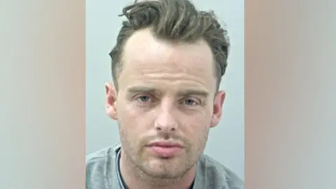 Lancashire Police James Ward's mugshot against a pale grey background. He has dark hair, long on top and swept back with a receding hairline and stubble. He is wearing a grey t-shirt.