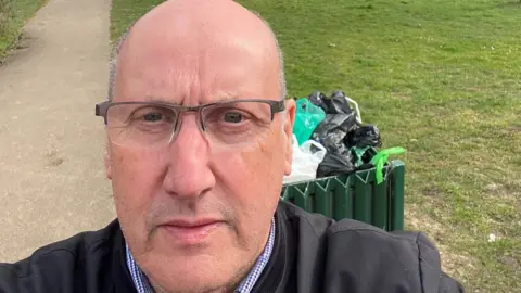 Doug is wearing a blue jacket and is posing behind a full bin in a park in Maidenhead. 