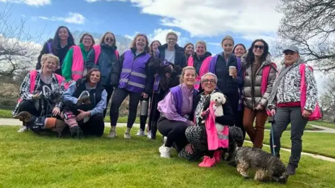 Leesa Collinge A group of women, wearing pink and purple hi-vis vests, stand outdoors with dogs.