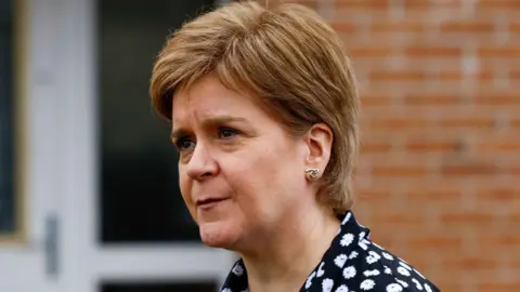 Getty Images Nicola Sturgeon standing outside her front door she is looking top the right and is wearing a black shirt with white flowers on it